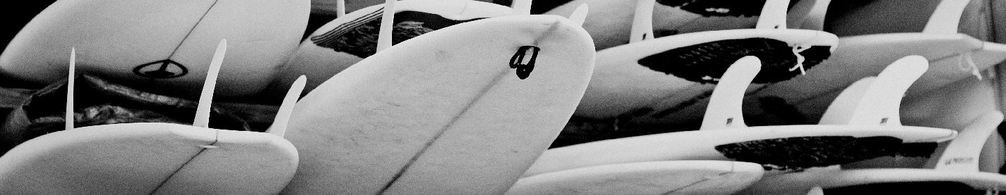 A black and white close-up of several surfboards stacked together, highlighting their fins and textured bottoms.